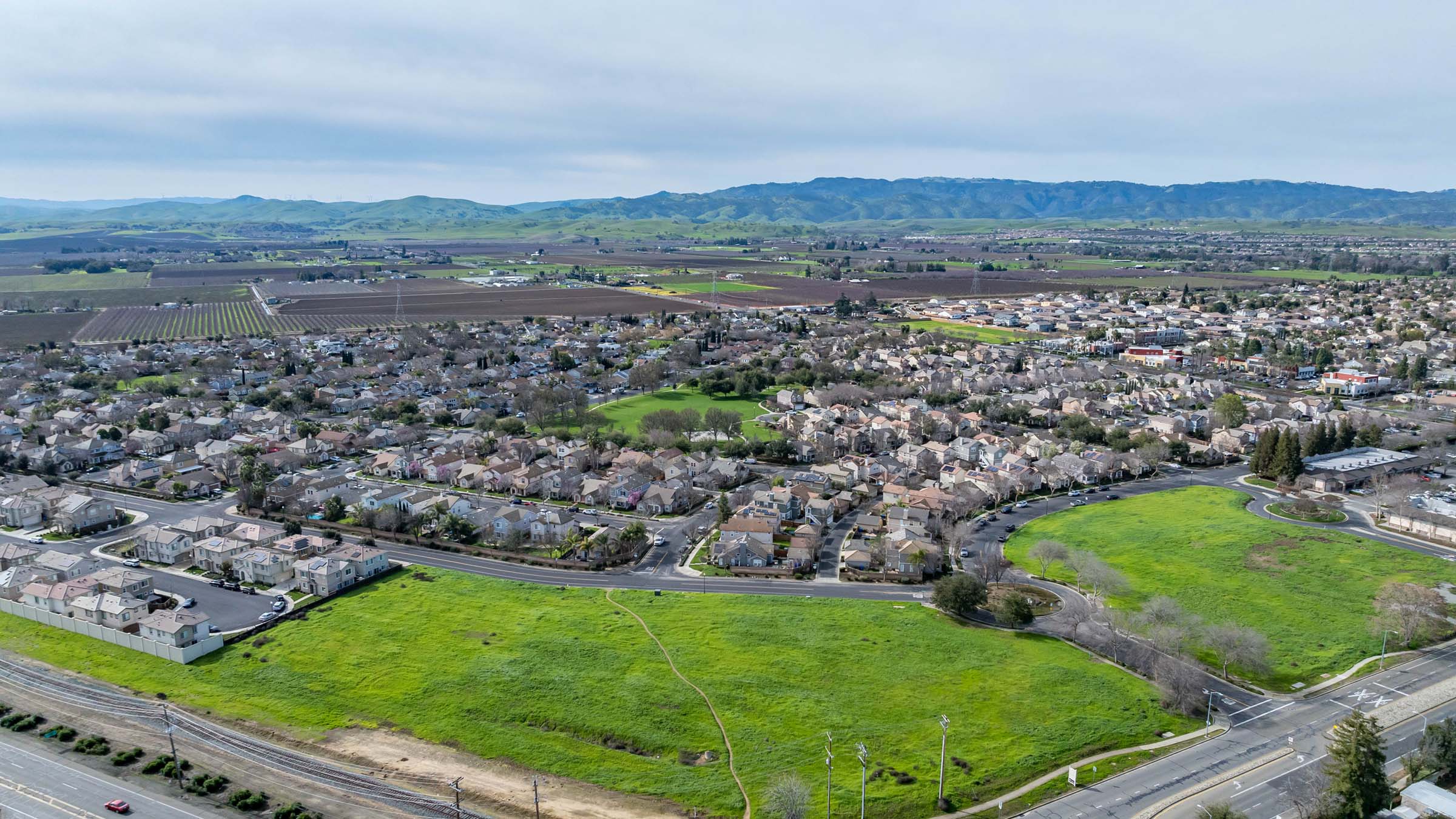 A drone-level view of a newly developed neighborhood in a suburban area in Northern California. Two large tracts of green, undeveloped land appear in the foreground. Multiple subdivisions containing single-family homes stretch into the background, where agricultural fields and mountains are visible on the horizon.