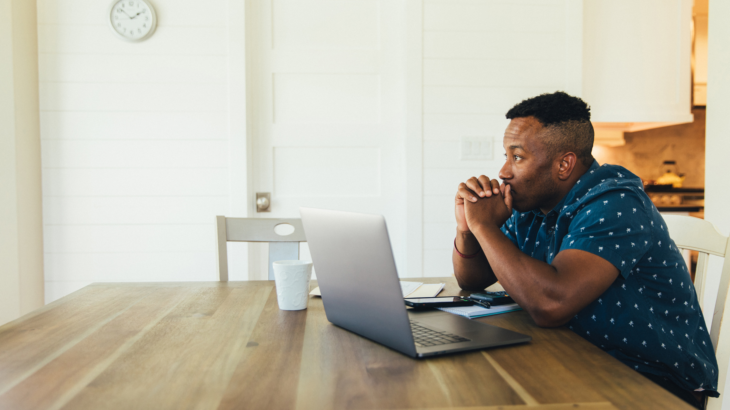 A young Black man sits at his kitchen table with a laptop computer open in front of him. He's looking into the distance with a worried or pensive expression on his face.