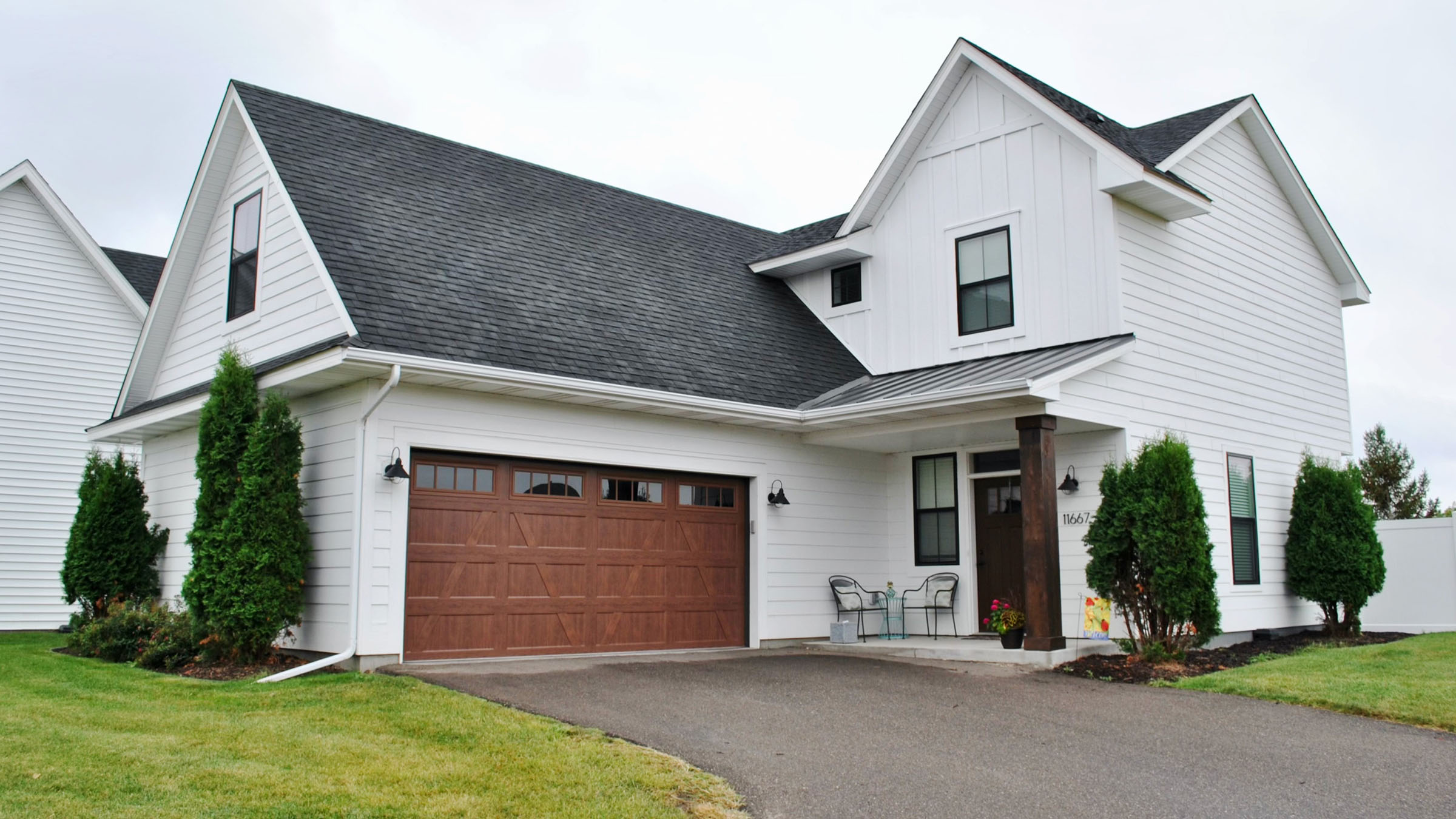 A tidy, two-story, newer construction home in a suburban neighborhood. The home has a farmhouse-style exterior with white siding, a black roof and trim, and a brown garage door.
