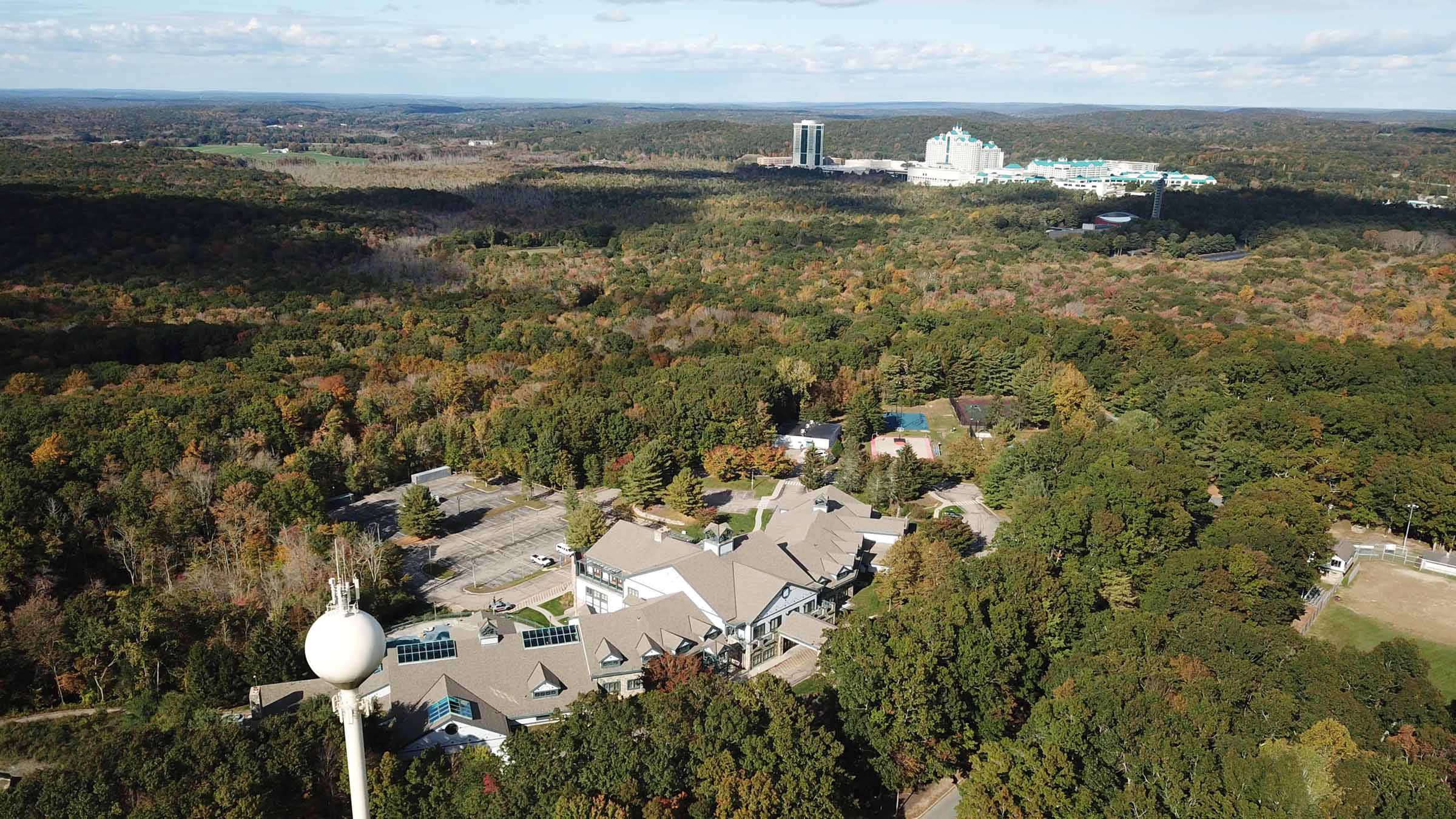 A daytime aerial view of the Mashantucket Pequot Tribal Nation reservation showing the tribal headquarters in the foreground and the tribe’s casino resort in the background. Both facilities are surrounded by hardwood forest that’s beginning to show autumn leaf colors.