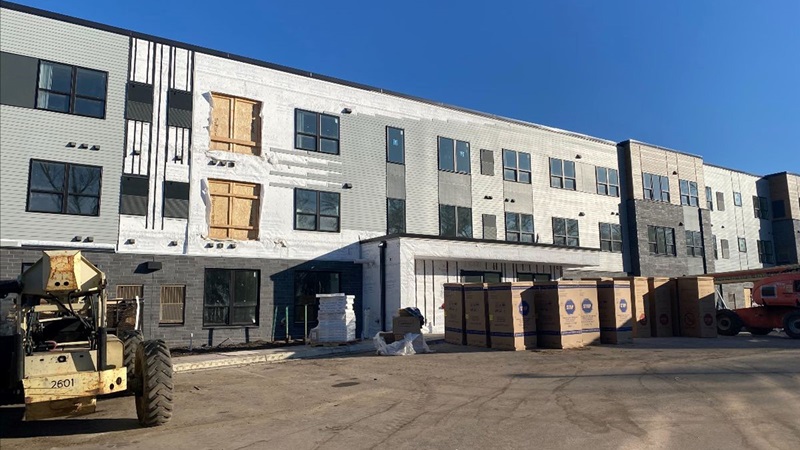 A three-story apartment building under construction, with the exterior mostly completed. In the parking area outside the building's entrance are construction machinery, including a small crane, and boxes of building materials. The day is clear and sunny.