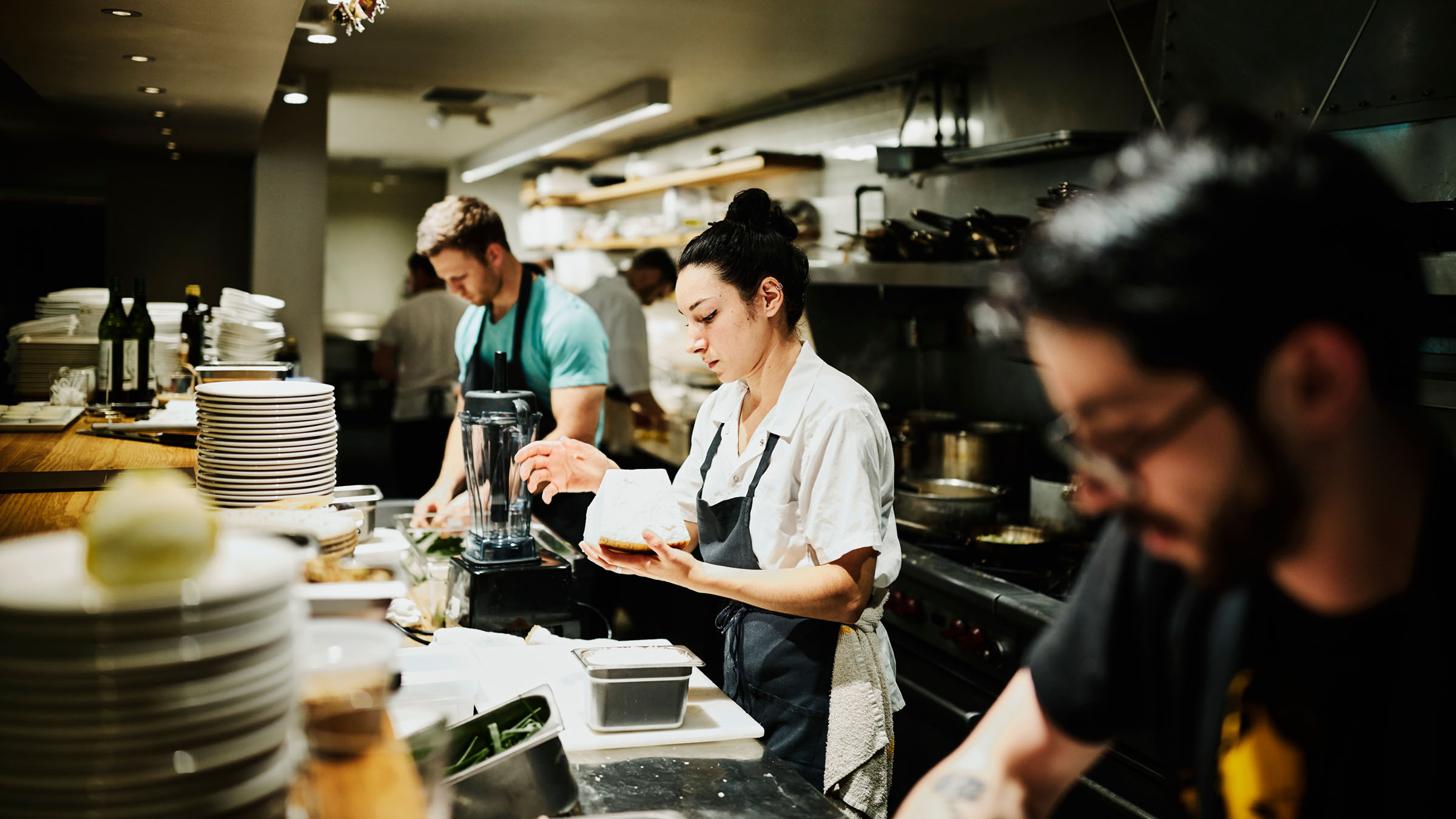 In the food-prepping station of a busy restaurant kitchen, a young female chef holds a large block of cheese to shred. She is flanked by two young male chefs who are absorbed in other food-prep tasks.