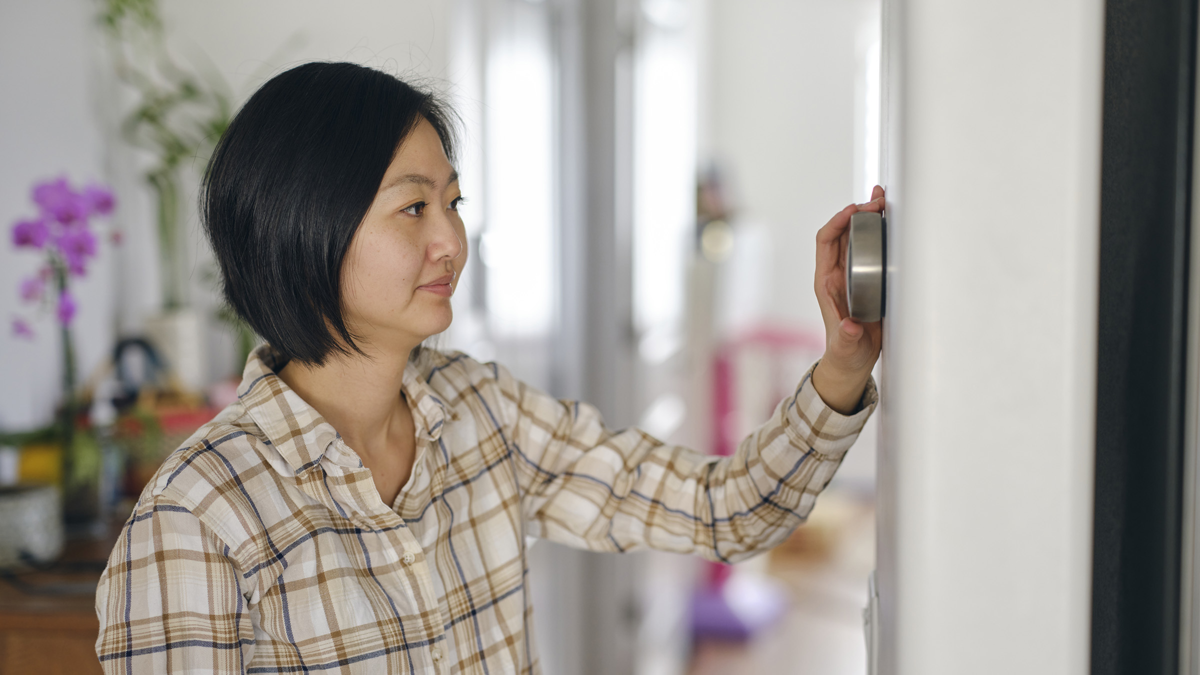 In an attractive, brightly lit home interior, a mature woman adjusts her thermostat. She is of Asian descent and is wearing a white button-down shirt with a brown and black plaid pattern. n