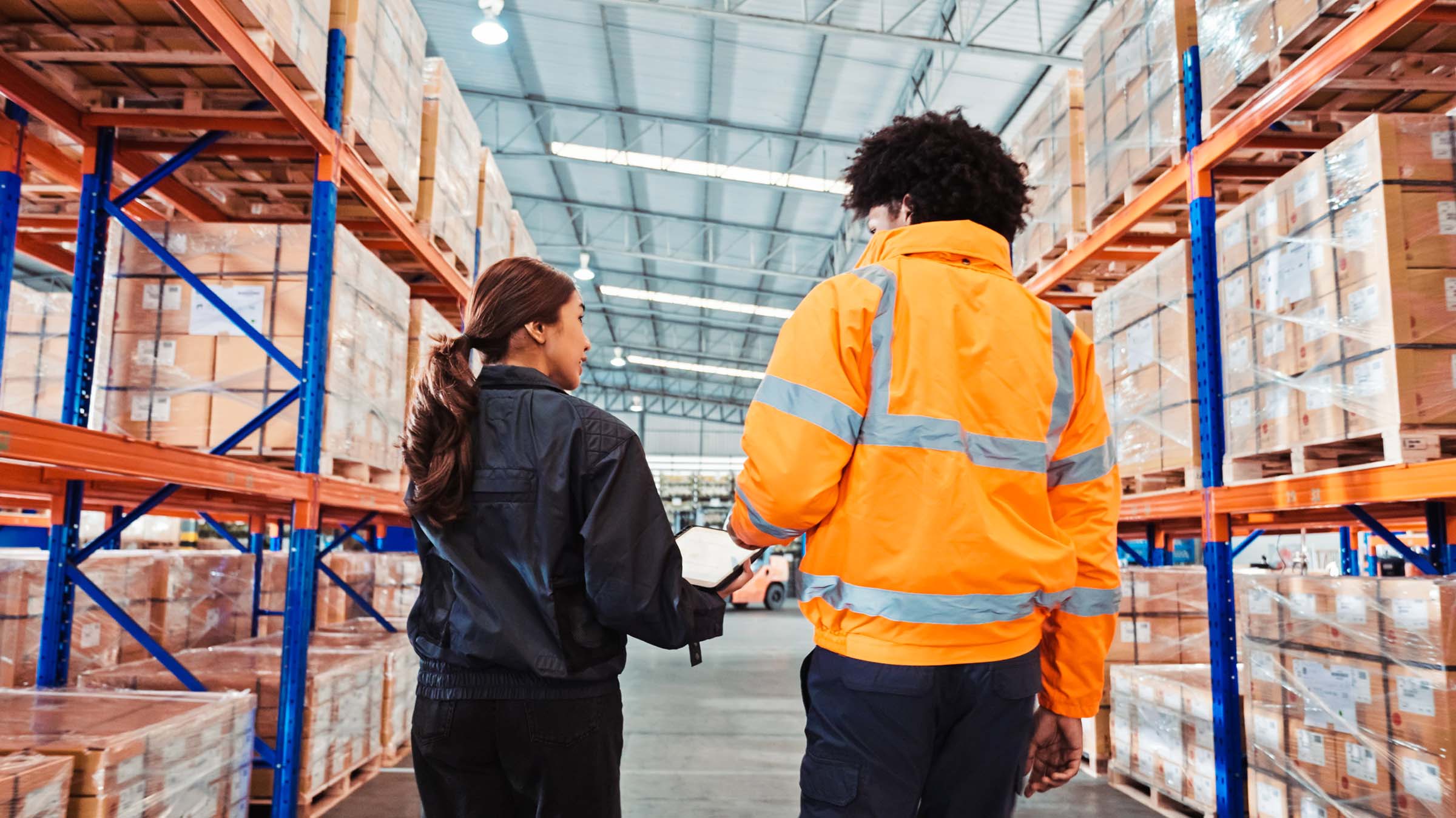 Viewed from behind inside a brightly lit warehouse, two workers--a young woman and a young man--walk down an aisle between stacks of shelving. The woman has long hair, a navy blue jacket, and black pants, and the man has short hair, an orange safety jacket with reflective stripes, and navy blue pants.