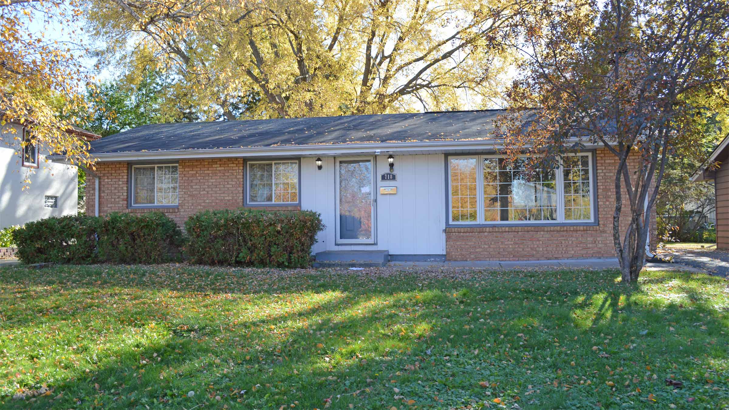 A small, mid-century, one-story house is pictured on a sunny, clear fall day. The house's exterior is a mixture of red brick and white siding.