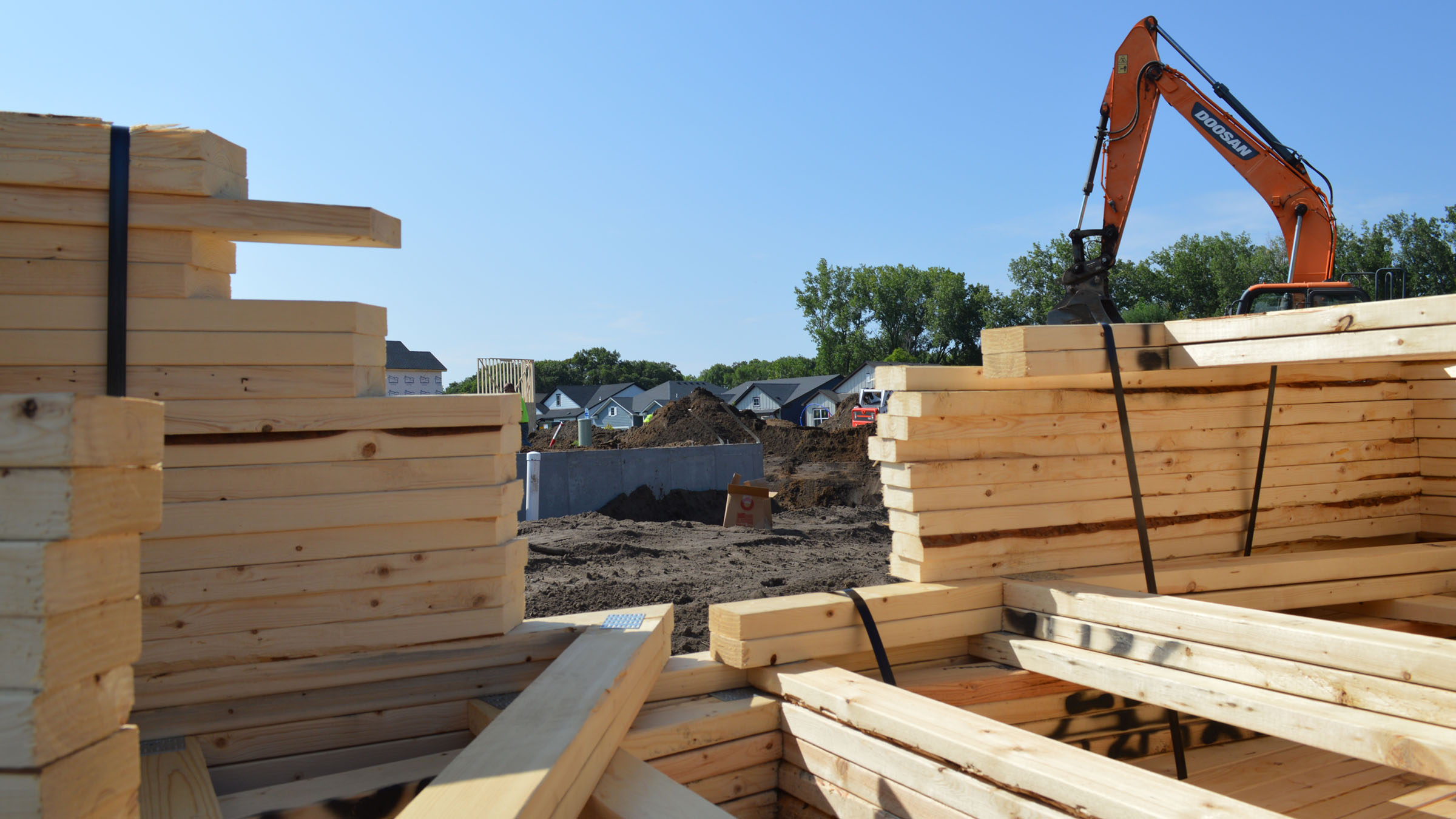At a single-family-home development site, pre-built wooden framing components are stacked in the foreground. Through a gap in the stacks, a partially constructed house is visible in the background. It's a clear summer day.