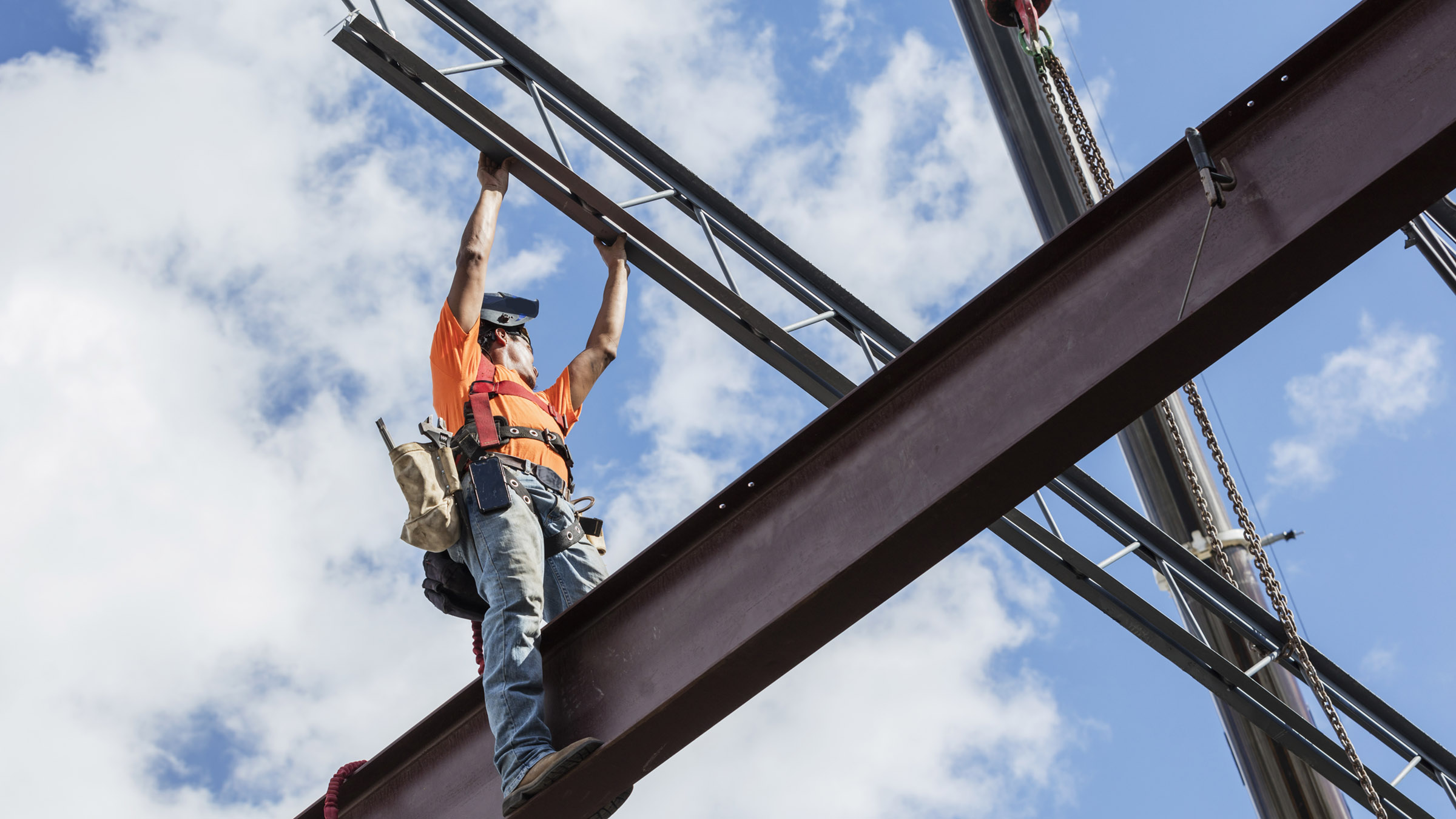 On a partly cloudy day, with the sky in the background, a male steel worker stands on a girder and reaches up for a roof joist or truss that is being lifted into place by a crane. He wears jeans, an orange tee shirt, and a safety harness, and has a welding helmet on his head.