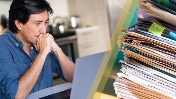 Photo on left side of image: A man in a denim-blue button-down shirt sits at his kitchen table with his chin in his folded hands, looking intently at his laptop screen. Photo on right side of image: Close-up of a tall, hastily stacked pile of paperwork, some of it with brightly colored Post-It flags and notes attached.