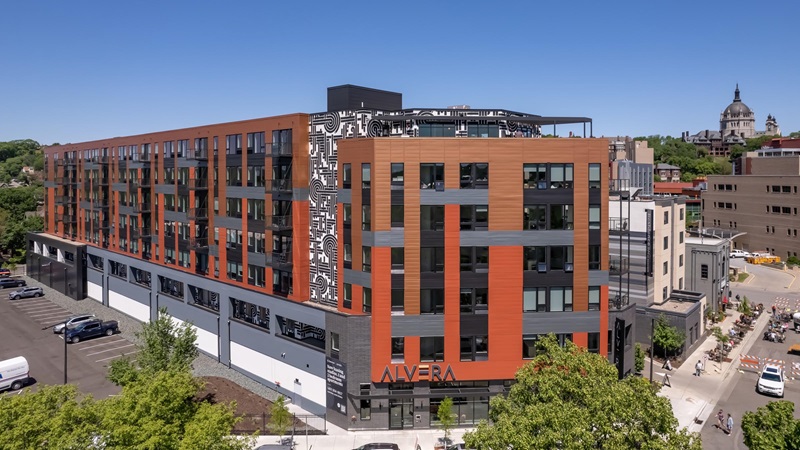 A crane- or drone-level view of Alvera Apartments, a modular housing building in St. Paul, on a clear summer day. The seven-story building has a modern exterior clad in red-orange, brown, and gray sheathing and features a black-and white mural along one wall.