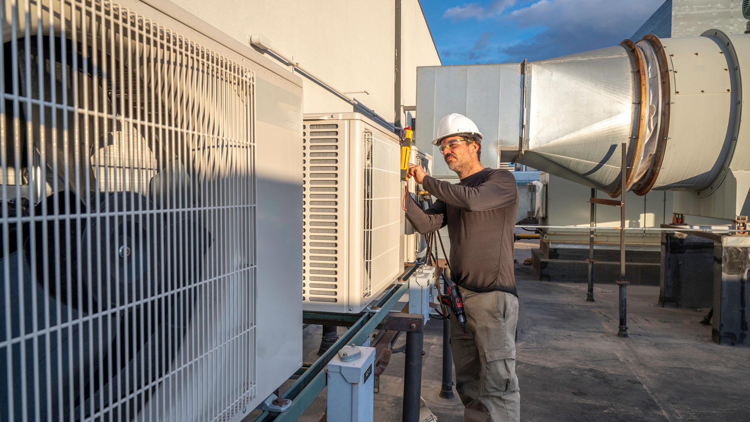 A male HVAC technician in a black long-sleeve tee shirt, gray pants, and white hard hat services commercial air conditioning equipment on the roof of an industrial building. A patch of partly cloudy sky is visible in the background.