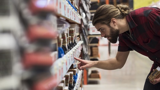 Inside a big-box home improvement store, a man in a short-sleeve plaid shirt bends down to compare the prices of boxes of hardware that are located on a low shelf. He is seen in profile, with the camera looking down the length of the store aisle.