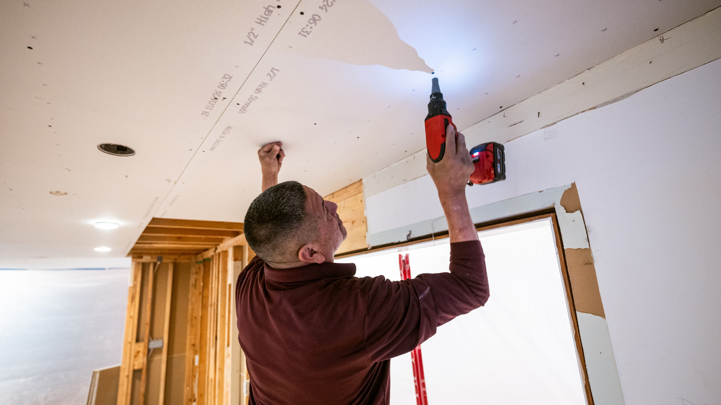 In a brightly lit residential kitchen that is partially stripped down to the studs, a drywall installer with his back to the camera uses a cordless drill to attach a drywall panel to the ceiling joists. He wears a long-sleeve, maroon-colored shirt and is using his left hand to brace the panel in place as he holds the drill in his right hand.