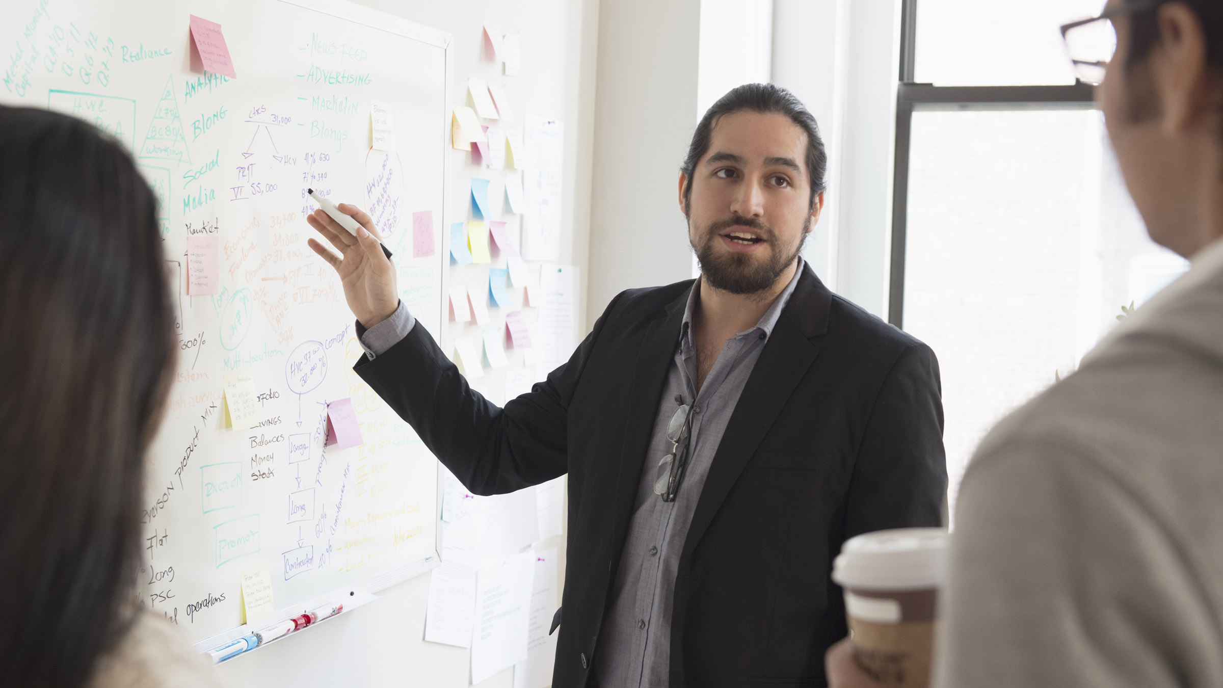 In a brightly lit office space, a man in a gray button-down shirt and black blazer stands at a white board and speaks to two colleagues, a man and a woman, who are partially visible from behind. The speaking man holds a marker close to the white board, which is covered with notes and colorful Post-Its, and looks at his male colleague as he speaks.