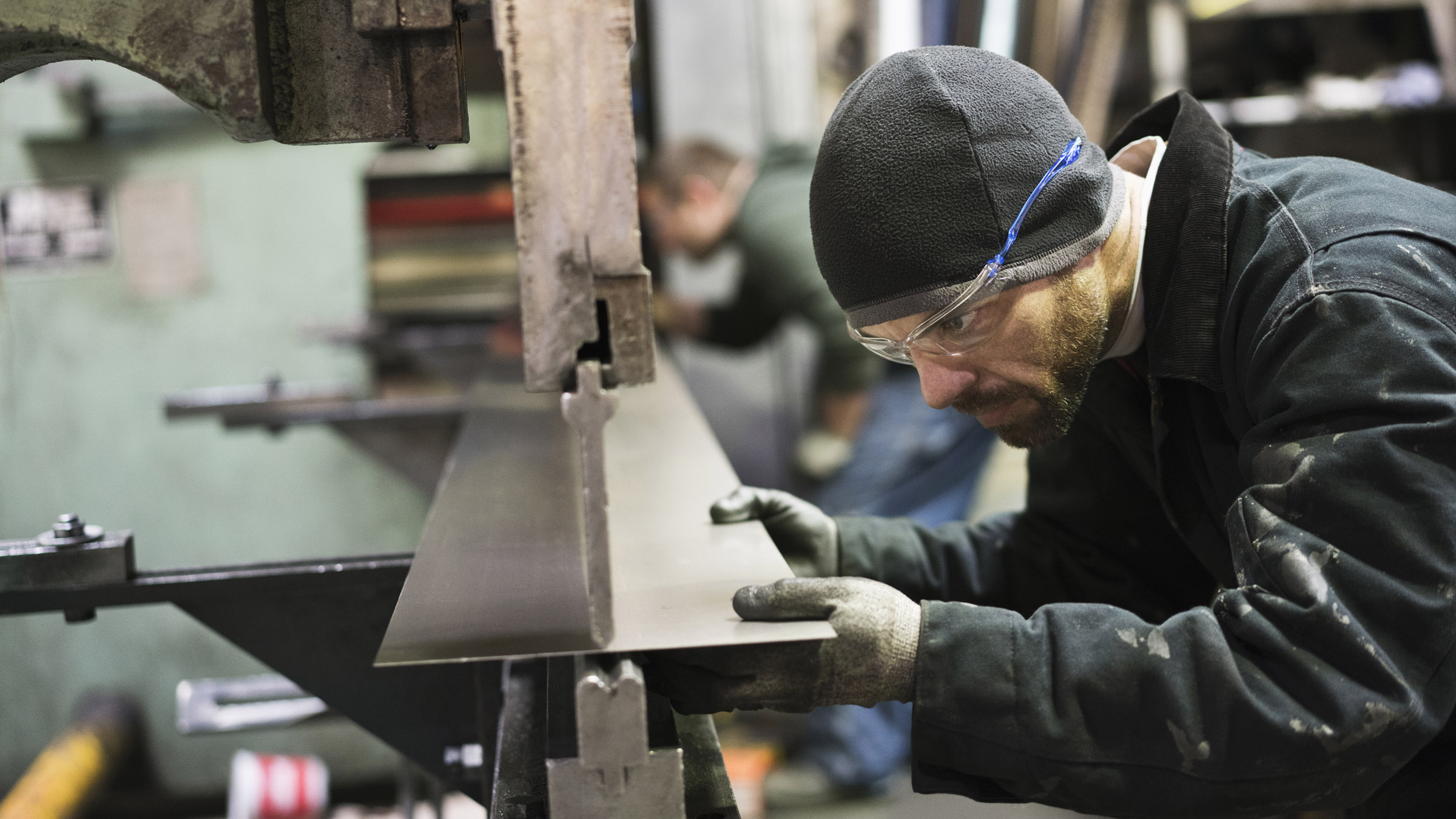 Inside a metal fabrication workshop, a male worker leans forward and holds a long, narrow sheet of steel in place on one end of a large metal press. The machine and the worker are seen in profile, with the worker wearing a black jacket, gray cap, gray protective gloves, and safety glasses. A second worker can be seen out of focus in the background, securing the other end of the steel panel.