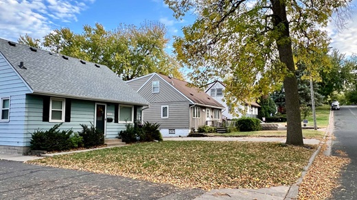 A view looking up a residential street in Columbia Heights, Minnesota, on a partly cloudy fall day. Three small Cape Cod-style homes can be seen: a light blue house in the foreground, a beige house in the middle, and a white house in the background.