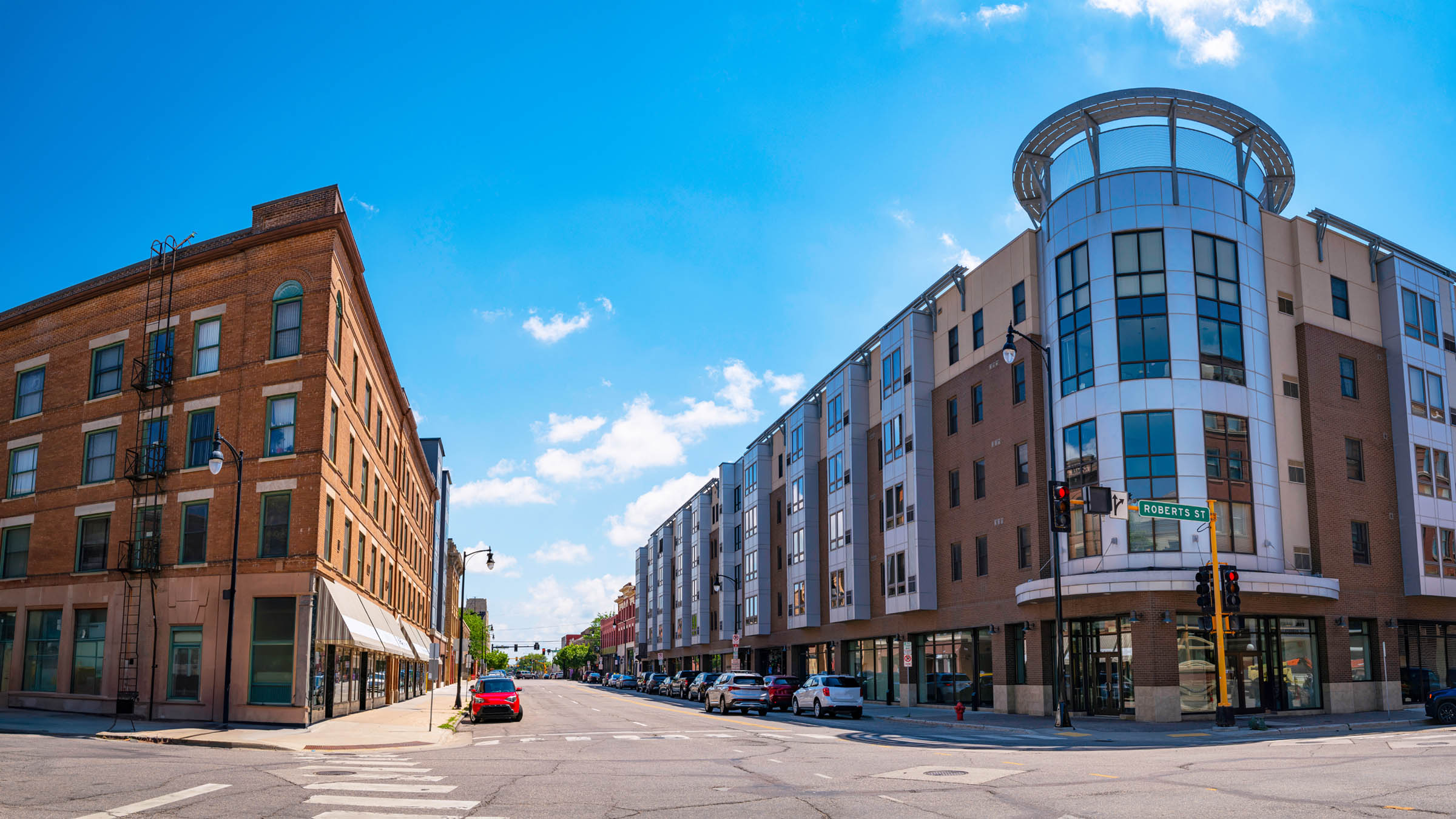 A street-level view of the intersection of First Avenue North and Roberts Street North in Fargo, North Dakota, looking east on a partly cloudy summer day. An older, four-story, brick building appears on the left and the newer-construction, five-story Cityscapes Plaza apartments appears on the right.