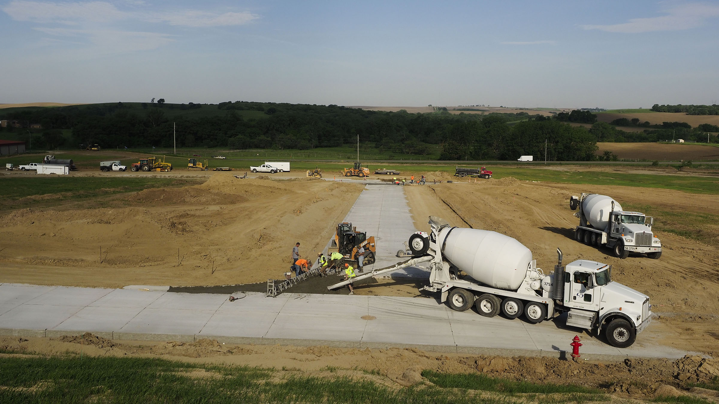 A high-angle view of a rural construction site on a sunny summer day. In the foreground are two white concrete-mixing trucks and a crew of several workers who are laying down the concrete for a segment of roadway. In the background are additional construction vehicles and green, rolling farmland.